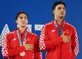 Two athletes from Croatia stand on a podium, wearing red and white checkered jackets and holding their medals with hands over their hearts.