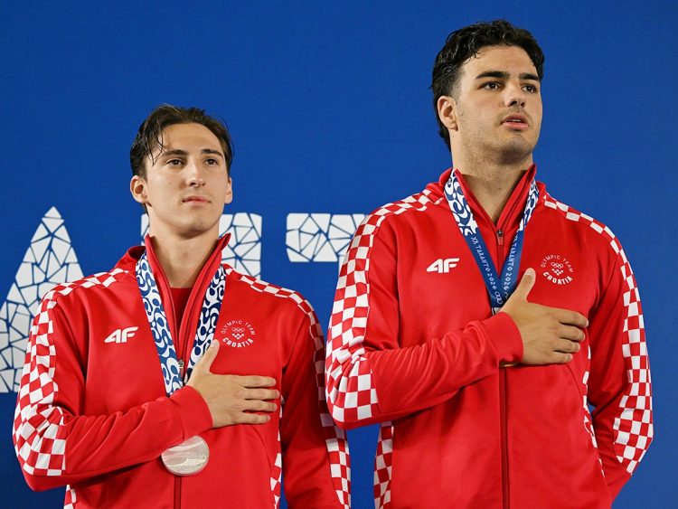 Two athletes from Croatia stand on a podium, wearing red and white checkered jackets and holding their medals with hands over their hearts.