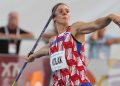 An athlete in a red and white uniform with blue shorts is mid-throw with a javelin during a competition.