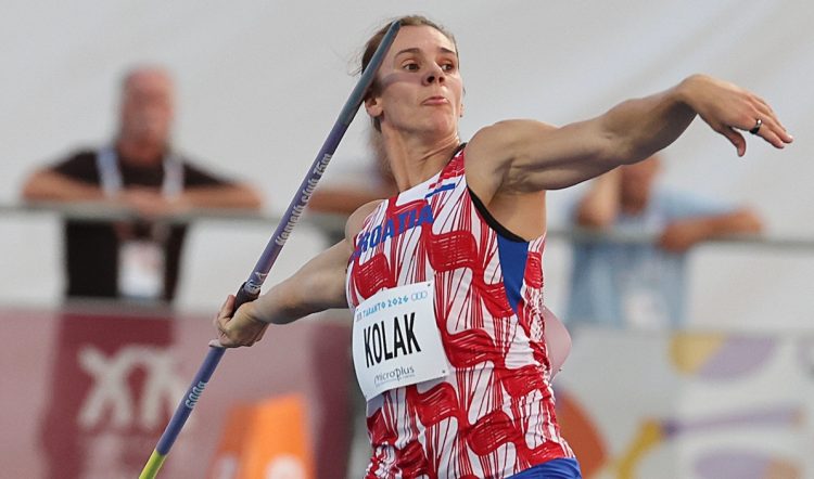 An athlete in a red and white uniform with blue shorts is mid-throw with a javelin during a competition.